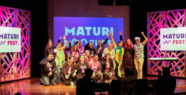 um grupo de pessoas reunida posando em frente a um telão que diz "MaturiFest"