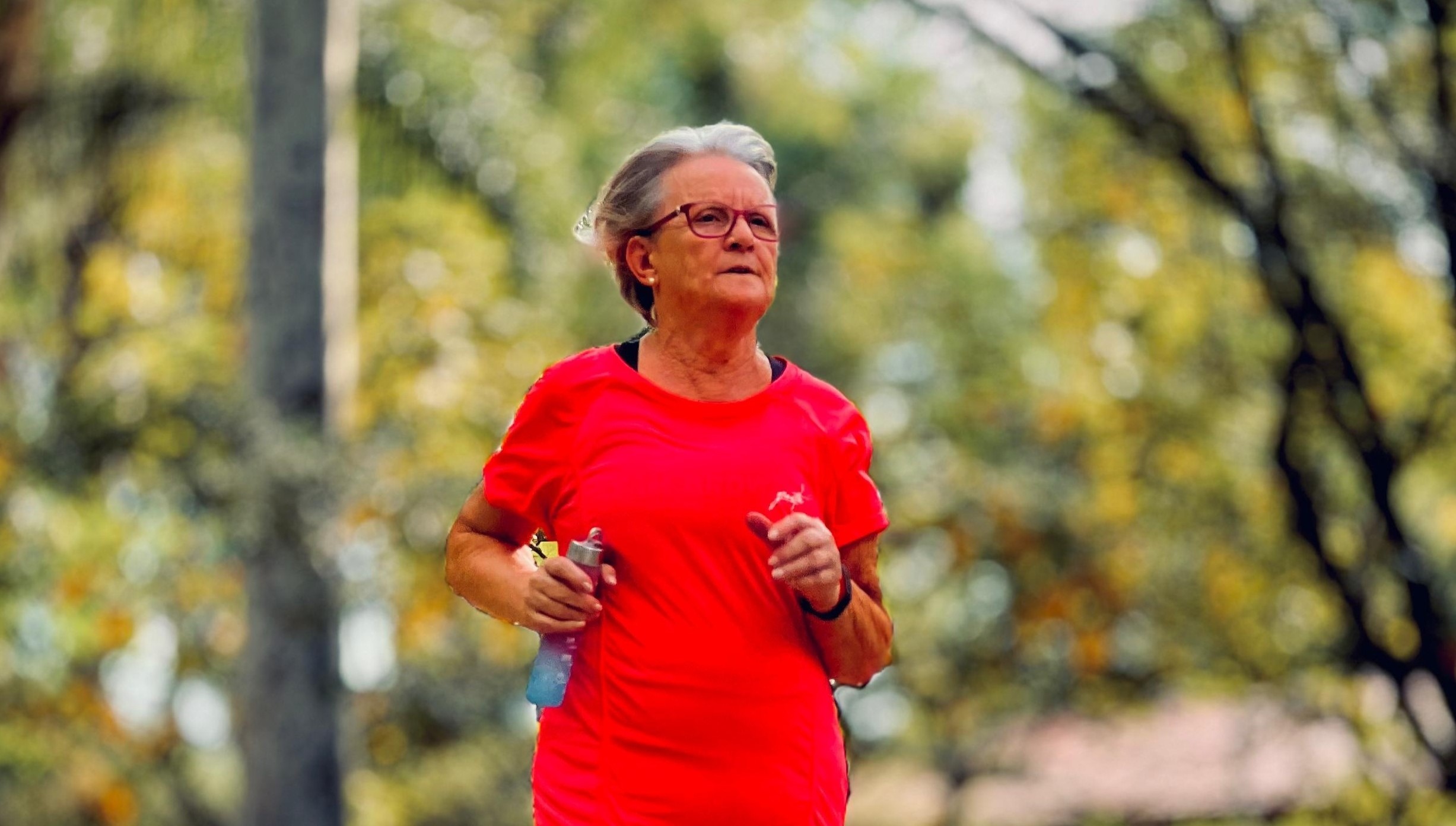 Uma senhora de pele branca, blusa vermelha correndo em um parque com árvores ao fundo.