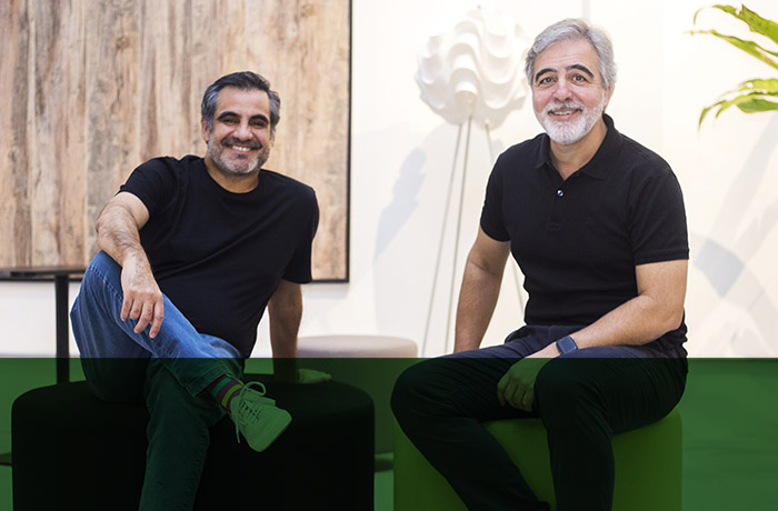 Dois homens brancos, com barba, usando camiseta preta, estão sentados sorrindo para uma foto